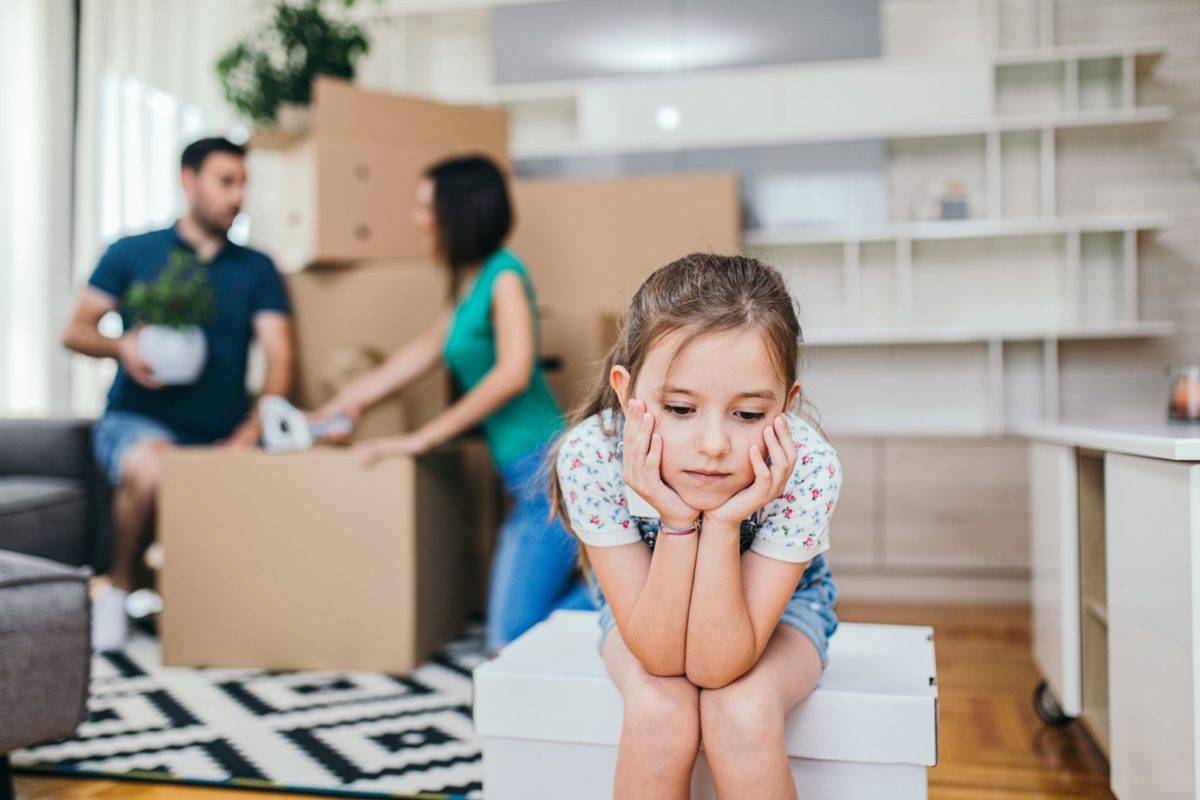 Little girl, sad about cross-country moving, sitting in front of her parents who are packing